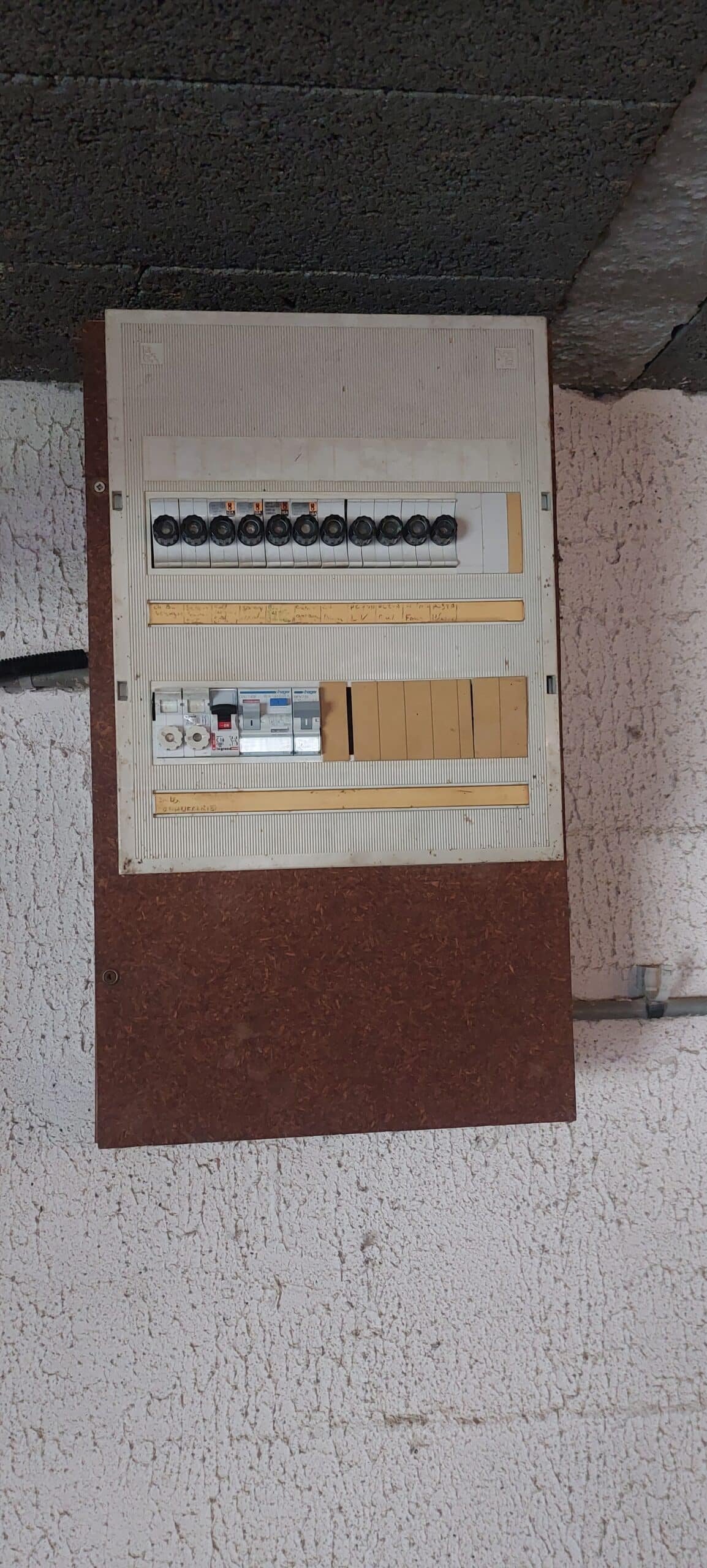 Tableau électrique ancien avec porte-fusibles et disjoncteurs Hager sur un mur crépi blanc.