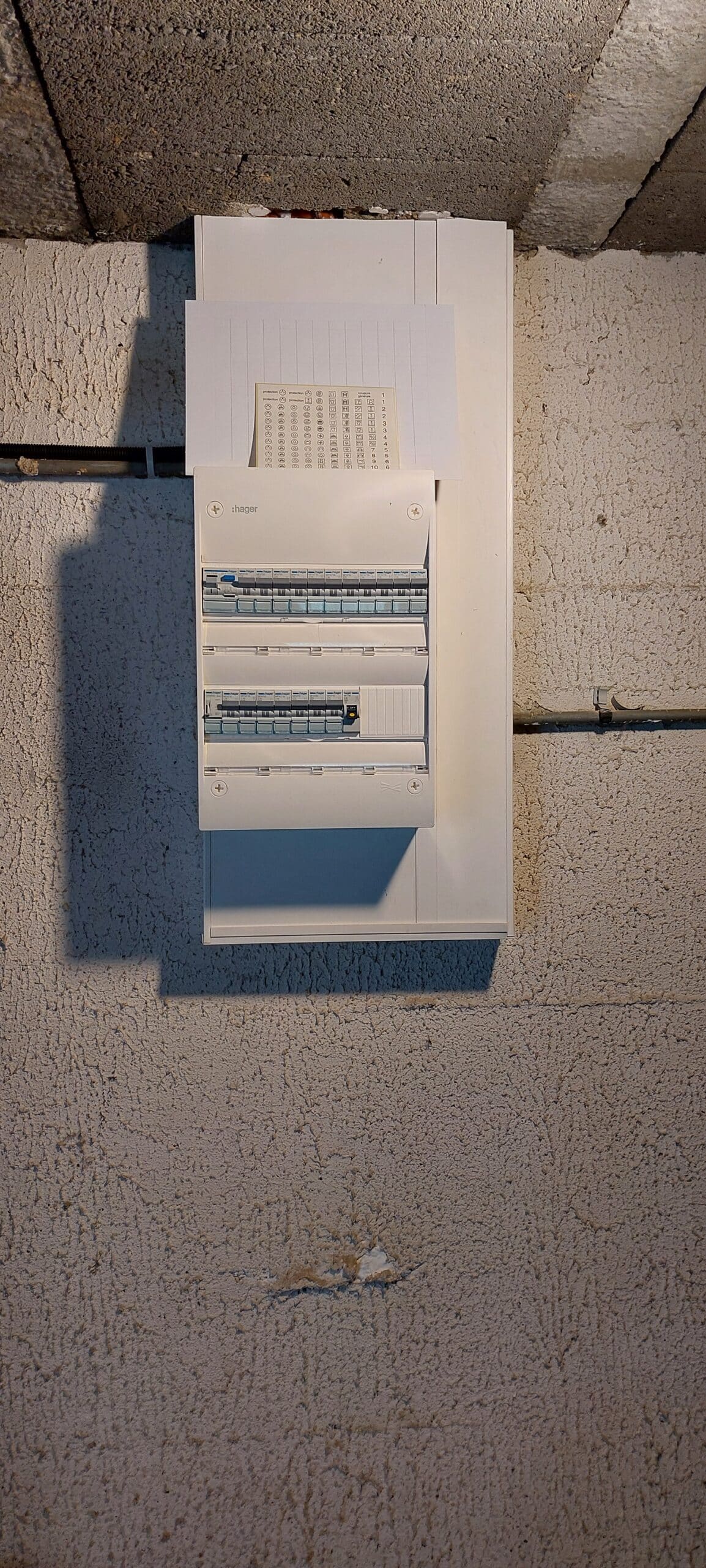 Tableau électrique Hager blanc avec rangées de disjoncteurs sur un mur de sous-sol en crépi.