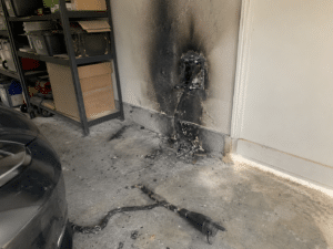 Mur de garage calciné et prise de recharge électrique fondue avec débris au sol suite à un court-circuit.